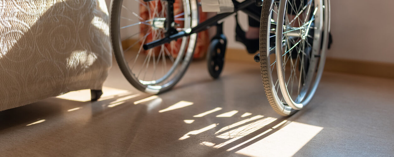 Detail of a wheelchair in front of a window and an elderly woman sitting, while enjoying the sun's rays