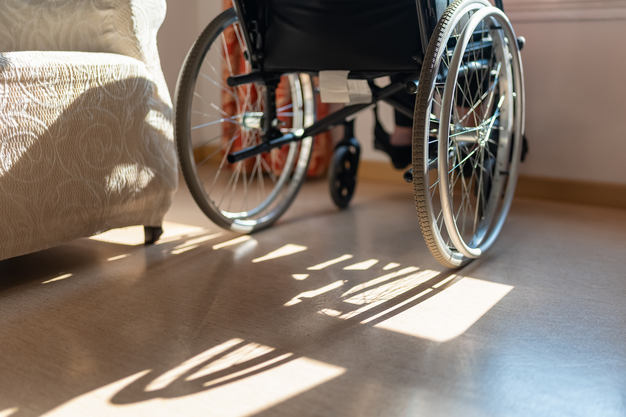 Detail of a wheelchair in front of a window and an elderly woman sitting, while enjoying the sun's rays