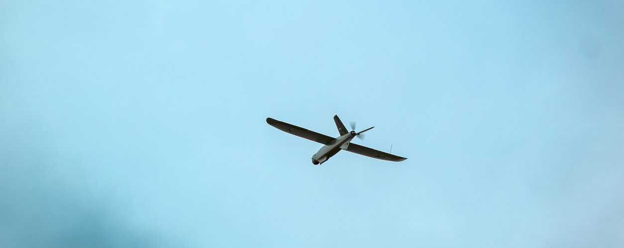 A drone flies against a bright blue sky with scattered clouds, surrounded by blurred green leaves in the foreground. aerial reconnaissance work.