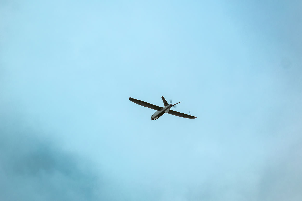 A drone flies against a bright blue sky with scattered clouds, surrounded by blurred green leaves in the foreground. aerial reconnaissance work.