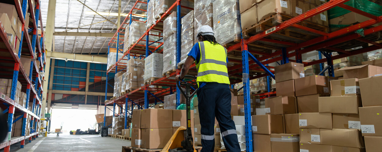 African American man worker using pallet truck for move cardboard boxes. Storehouse employee in uniform working and unloading goods in warehouse. Logistics, Distribution Center concept.