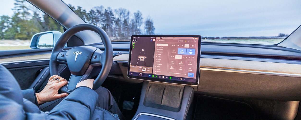 Sweden. Uppsala. 02.20.2025. Close-up view of driver holding steering wheel inside Tesla Model 3 electric car with touchscreen displaying vehicle controls.