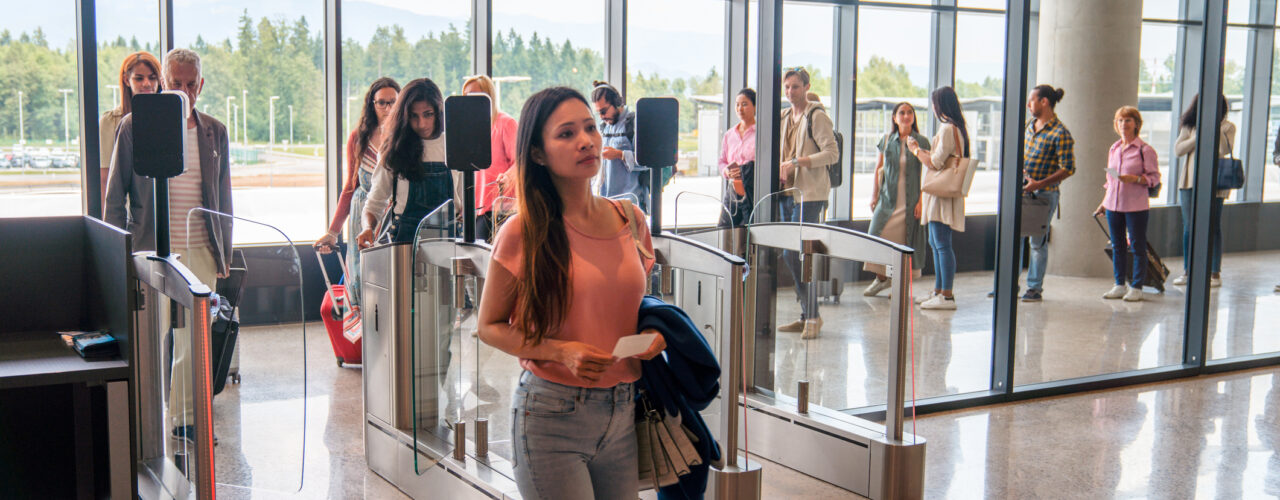 Multi ethnic group of female and male travelers queueing at the e-gates of an airport, a crowd behind the beautiful mid adult Asian female who already passed through, waiting to self check the boarding pass. 3/4 length image of the Asian woman, wide angle shot.